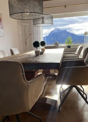 A modern dining area with a wooden table and chairs, potted plants, and a view of mountains.
