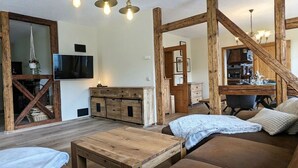 Un salon chaleureux avec un canapé en bois, une table basse et un téléviseur fixé au mur. Un escalier mène à un niveau supérieur où l’on aperçoit une cuisine.