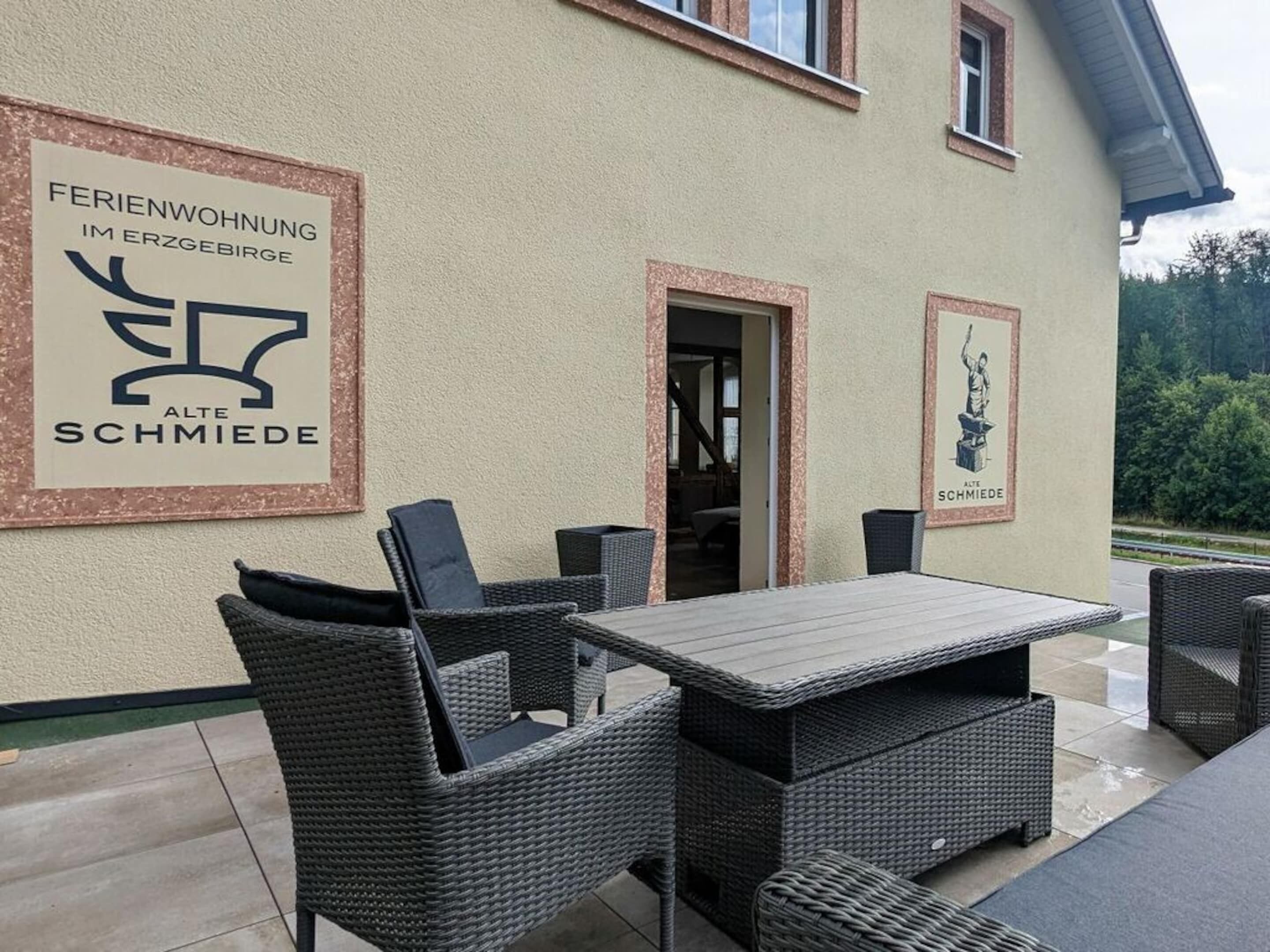 An outdoor patio area with wicker furniture and a table, a sign reading "Feriendorfung im Erzgebirge Alte Schmiede", and a framed picture of a person.