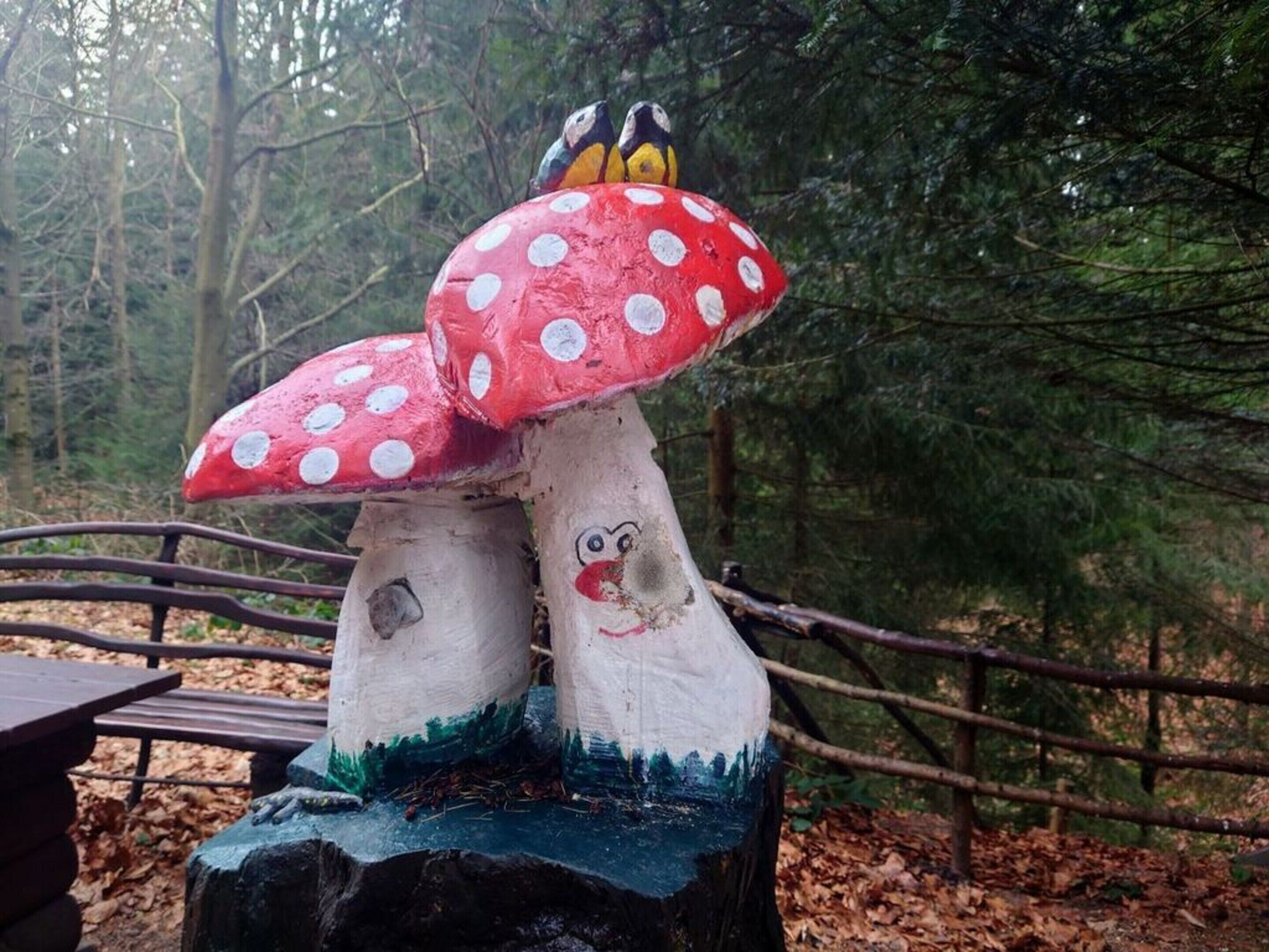 Una escultura de madera de dos hongos con rostros pintados y lunares en sus sombreros, sobre un fondo de bosque.