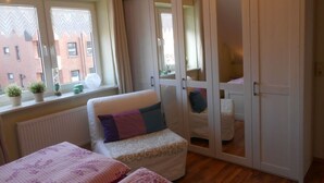 Een slaapkamer met een bed, een stoel, een kledingkast en een raam met planten.