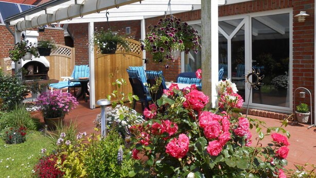 Een overdekt terras met buitenmeubilair, een huis van baksteen en een tuin met verschillende bloemen.