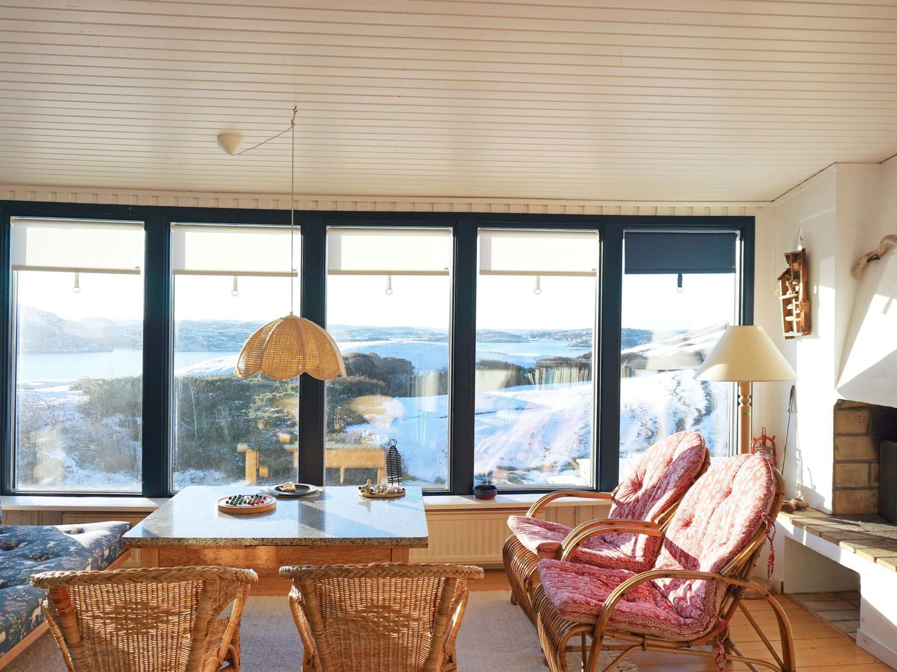 Ein Zimmer mit großem Fenster und Blick auf eine reizvolle Landschaft, einem Tisch mit dekorativen Gegenständen, Korbstühlen und einer Lampe.