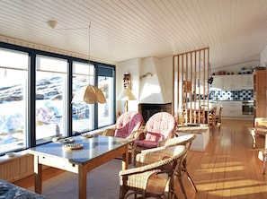 A cozy living room with a fireplace, wicker chairs, a dining table, and large windows offering a view of the outdoors. - 4 Star Holiday Home in Bovallstrand (Bovallstrand)