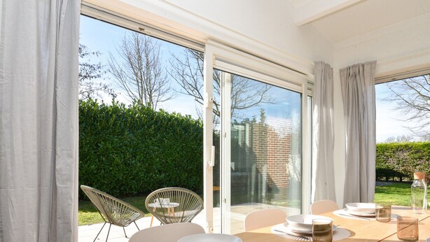 A dining area with a wooden table set for four, surrounded by chairs, with a view of a hedge and a brick wall through large glass doors. - Your Coastal Hideaway (Kamperland)