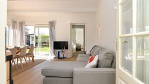 A modern living room with a grey sectional sofa, a dining area with a table and chairs, a flat-screen TV, and a large window with curtains. - Your Coastal Hideaway (Kamperland)