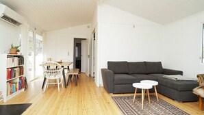 A spacious living room with a grey sofa, a small round table, and a wooden floor. There is a bookshelf filled with books and a potted plant on the left. - 5 Person Holiday Home in Melby (Melby)