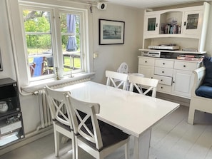 A kitchen with a white table and chairs, a radiator, a window with a view of greenery, and a cabinet with books and a Scrabble box. - 4 Star Holiday Home in Torhamn (Torhamn)