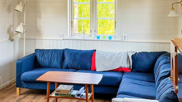 A room with a blue sofa, a wooden coffee table with books, a window with blinds, and a lamp. - 4 Star Holiday Home in Torhamn (Torhamn)