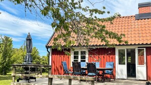 Ett trädäck med ett bord och stolar, en täckt uteplats och en röd byggnad med ett tak täckt av tegelpannor.