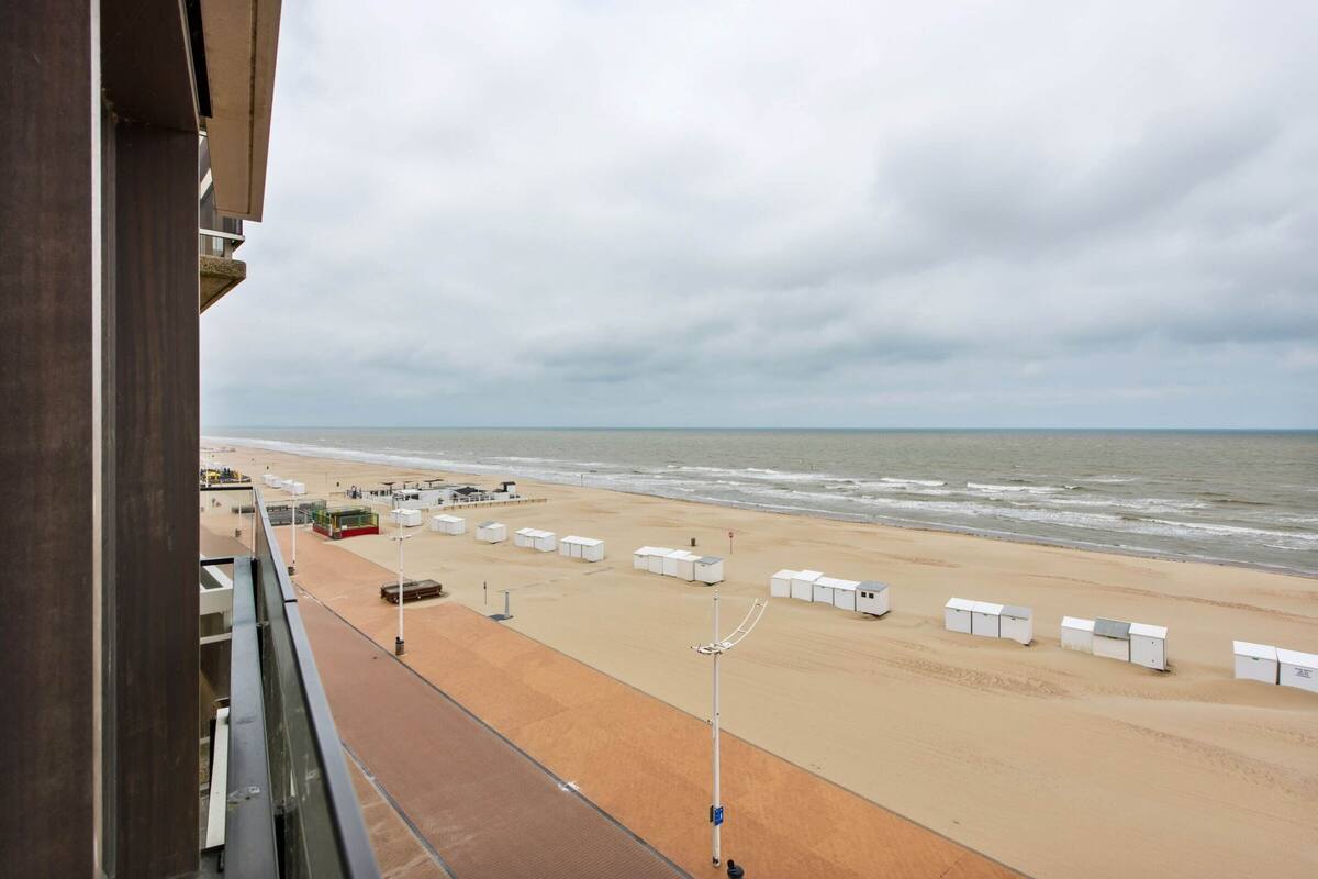 A beach view from a balcony with a sandy beach, a row of white beach huts, and a cloudy sky.