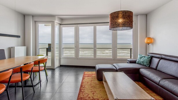 Un moderno salón con un ventanal grande con vistas al mar, una mesa de comedor de madera con sillas anaranjadas, un sofá de cuero oscuro y una lámpara colgante.