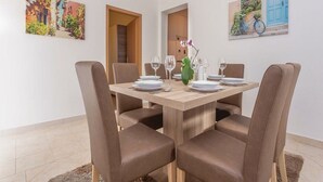 A dining area with a wooden table set for four, surrounded by brown upholstered chairs. There are two paintings on the walls, one depicting a colorful street scene and the other a blue door with a bicycle. - Apartment Niko - Two Bedroom Apartment With Swimming Pool (Sibenik)