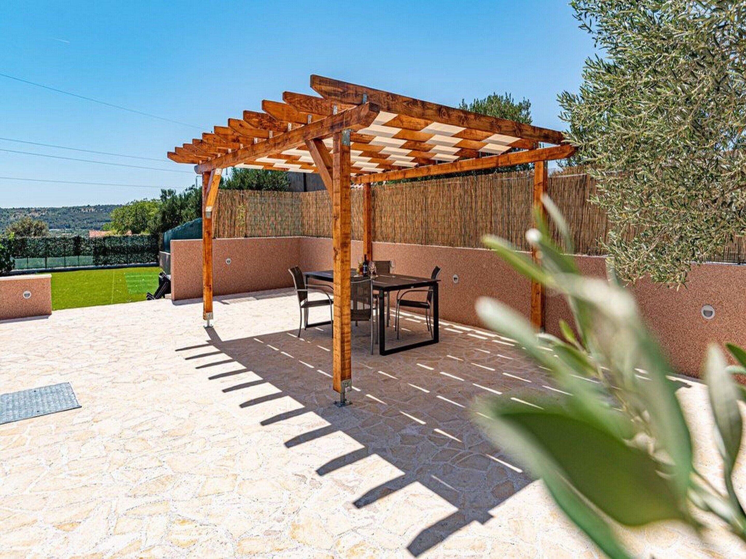 A covered outdoor patio with a pergola, a table and chairs, and a view of a green field.