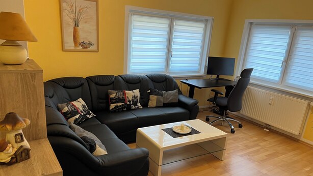 Un salon moderne avec un canapé en cuir noir, une table basse blanche et un fauteuil de bureau noir. Un tableau encadré est accroché au mur, et une lampe se trouve sur une petite table en bois.