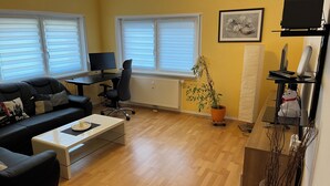 Un salon comprenant un canapé, une table basse et un bureau avec un ordinateur. Il y a deux fenêtres équipées de stores, un radiateur et une plante en pot.