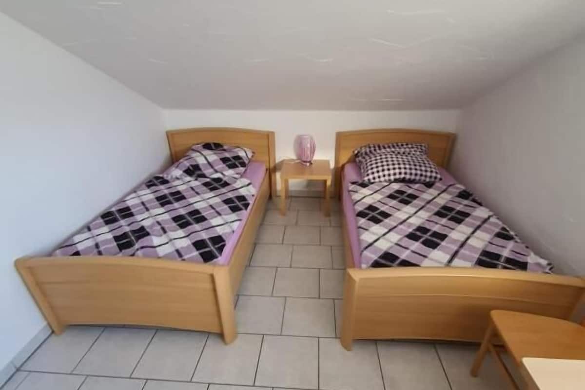 A small, clean room with two wooden beds, a small table, and a tiled floor.