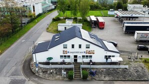 A white building with a sign reading "ERBACHER HOF" and a restaurant sign. There are parked trucks and a road in the background. - Erbacher Hof  (Homburg)