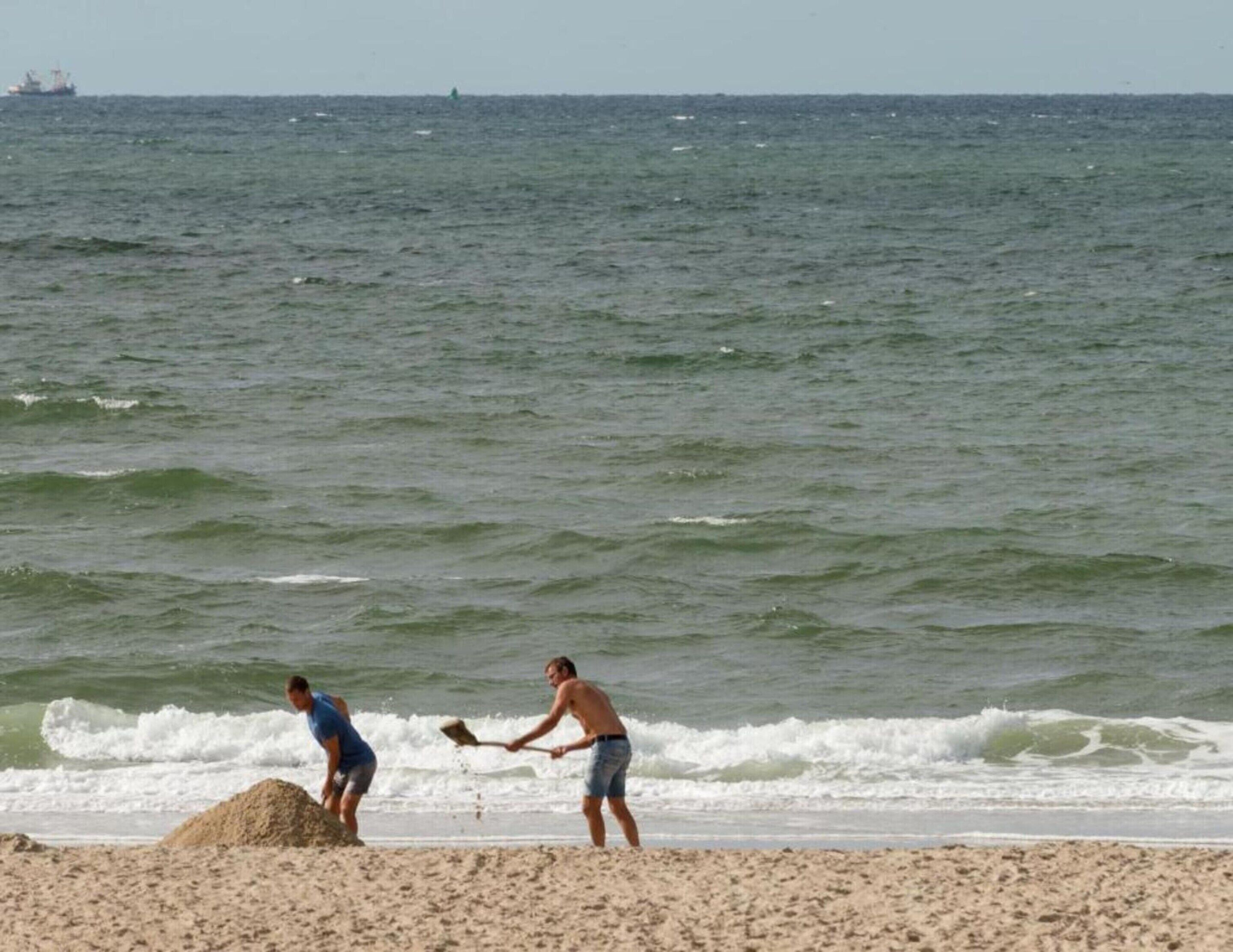 Two people on a beach near the ocean, one shoveling sand and the other holding a shovel.