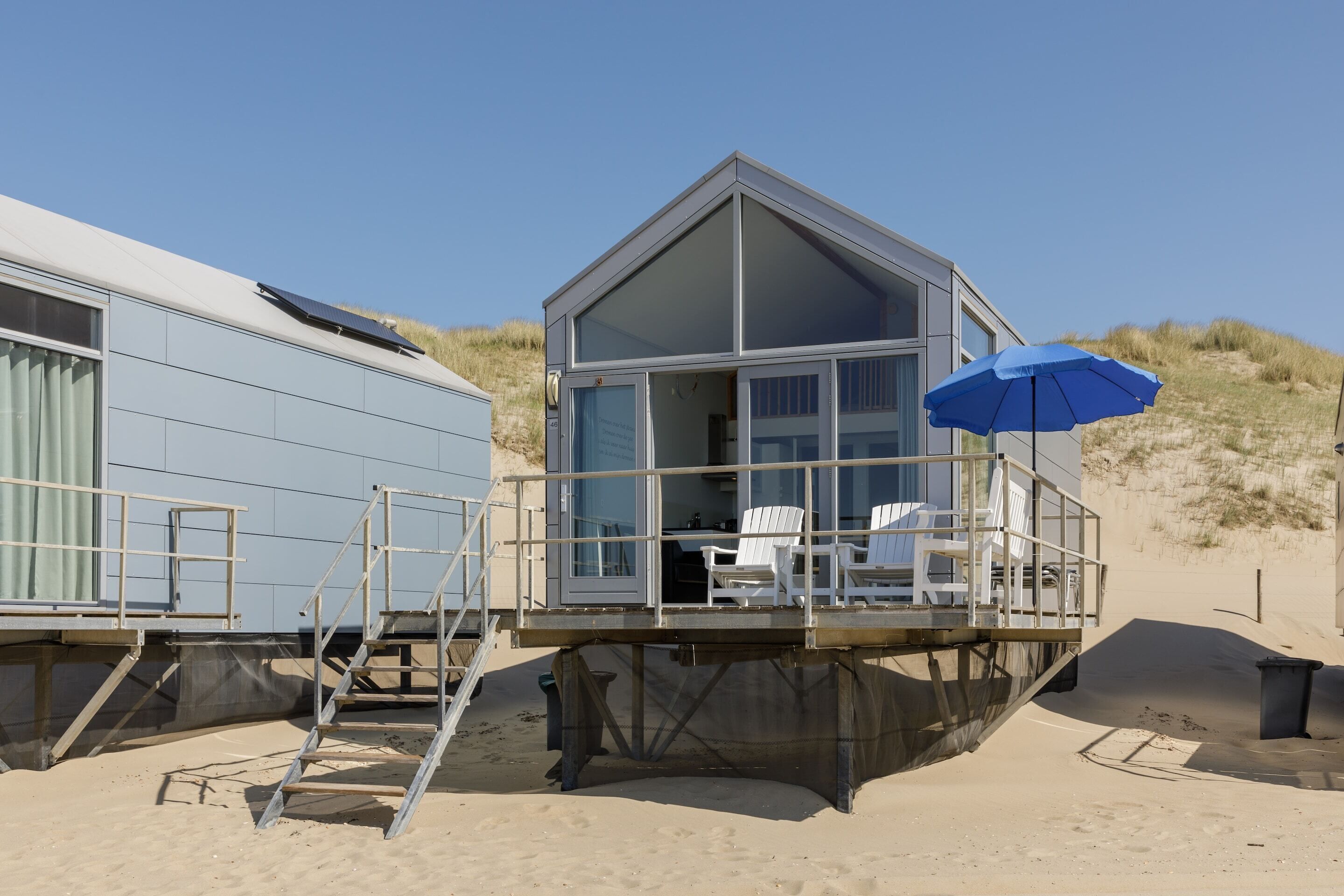 A beach house with a deck, a blue umbrella, and a white chair.