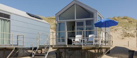 A beach house with a deck, a blue umbrella, and a white chair.