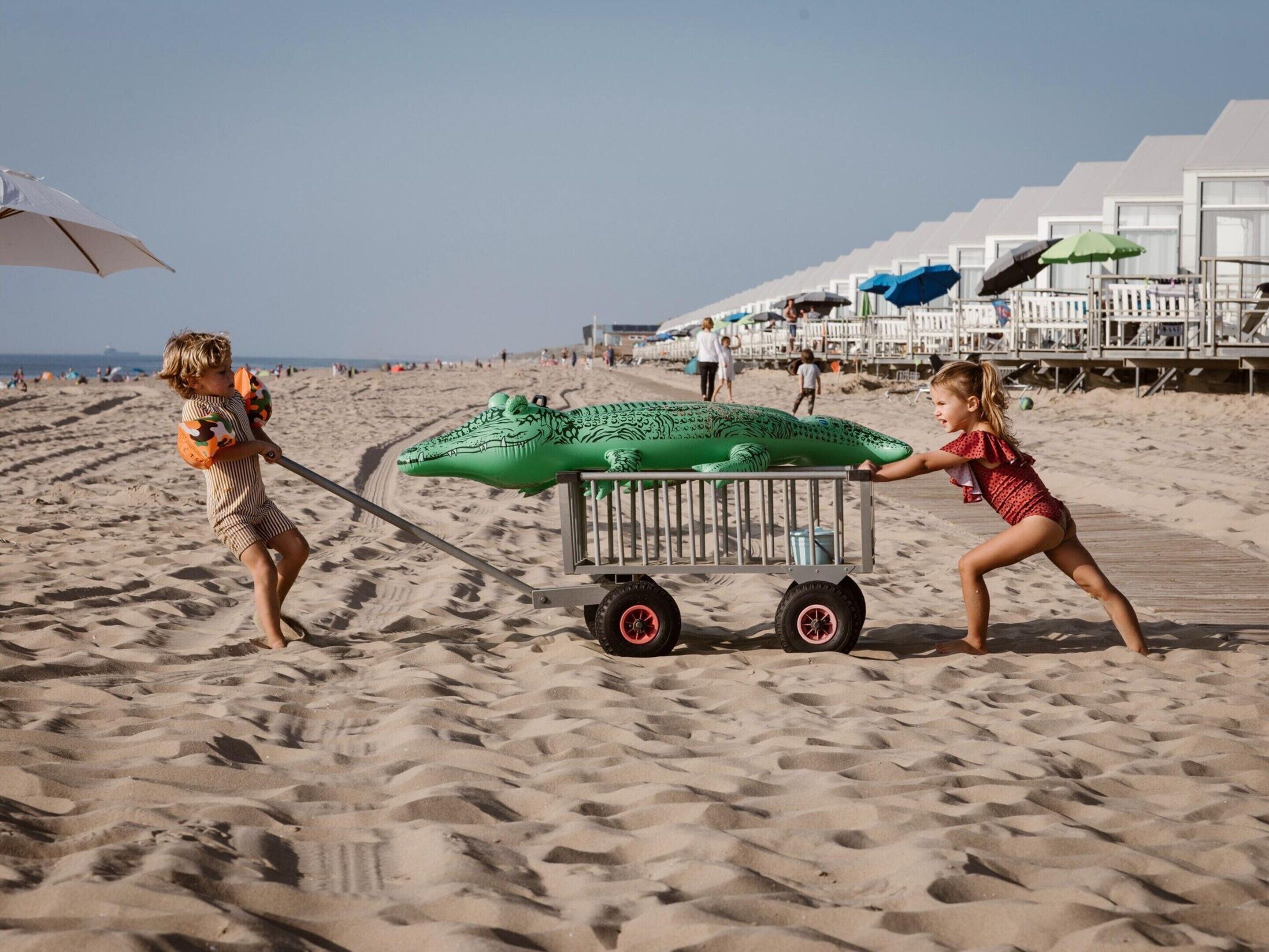 Escena de playa con un cocodrilo inflable verde que dos niños jalan.