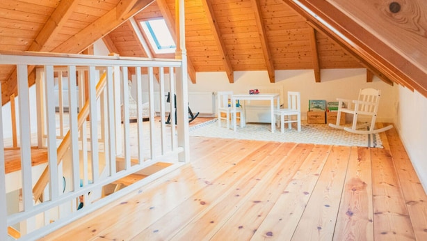 Um quarto de sótão espaçoso com teto de madeira inclinado, corrimão branco, uma pequena mesa com cadeiras e vista do céu através de um claraboia.