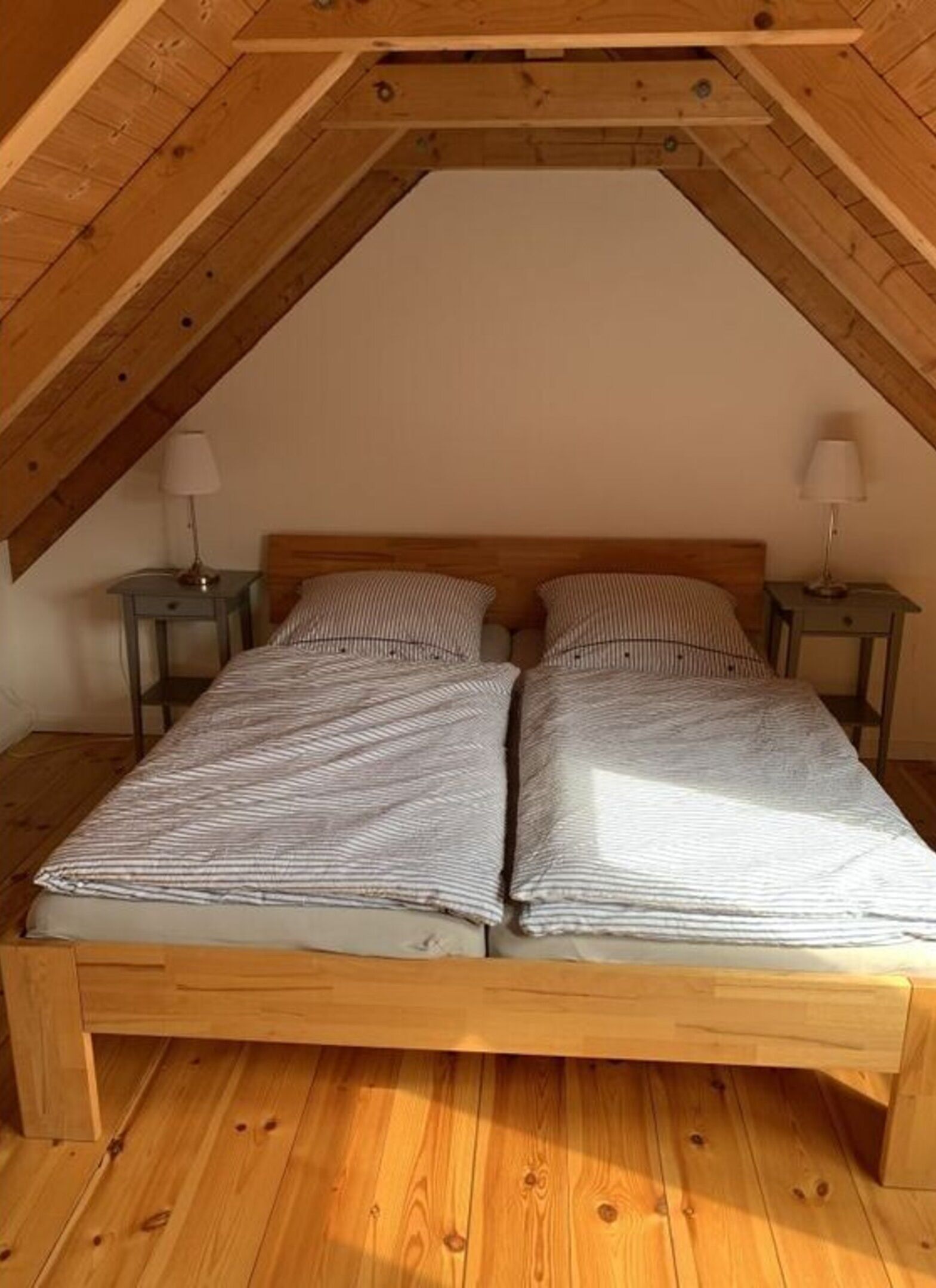 Una stanza con soffitto a falde, pavimento in legno e un letto con biancheria a righe. Sono presenti due comodini con lampade.