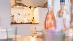 Uma cozinha com armários brancos, coifa de aço inoxidável e painel de azulejos. Há garrafas na bancada e um copo na mesa.