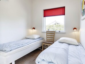 A bedroom with two single beds, a wooden chair, a window with a red blind, and two wall-mounted lamps.