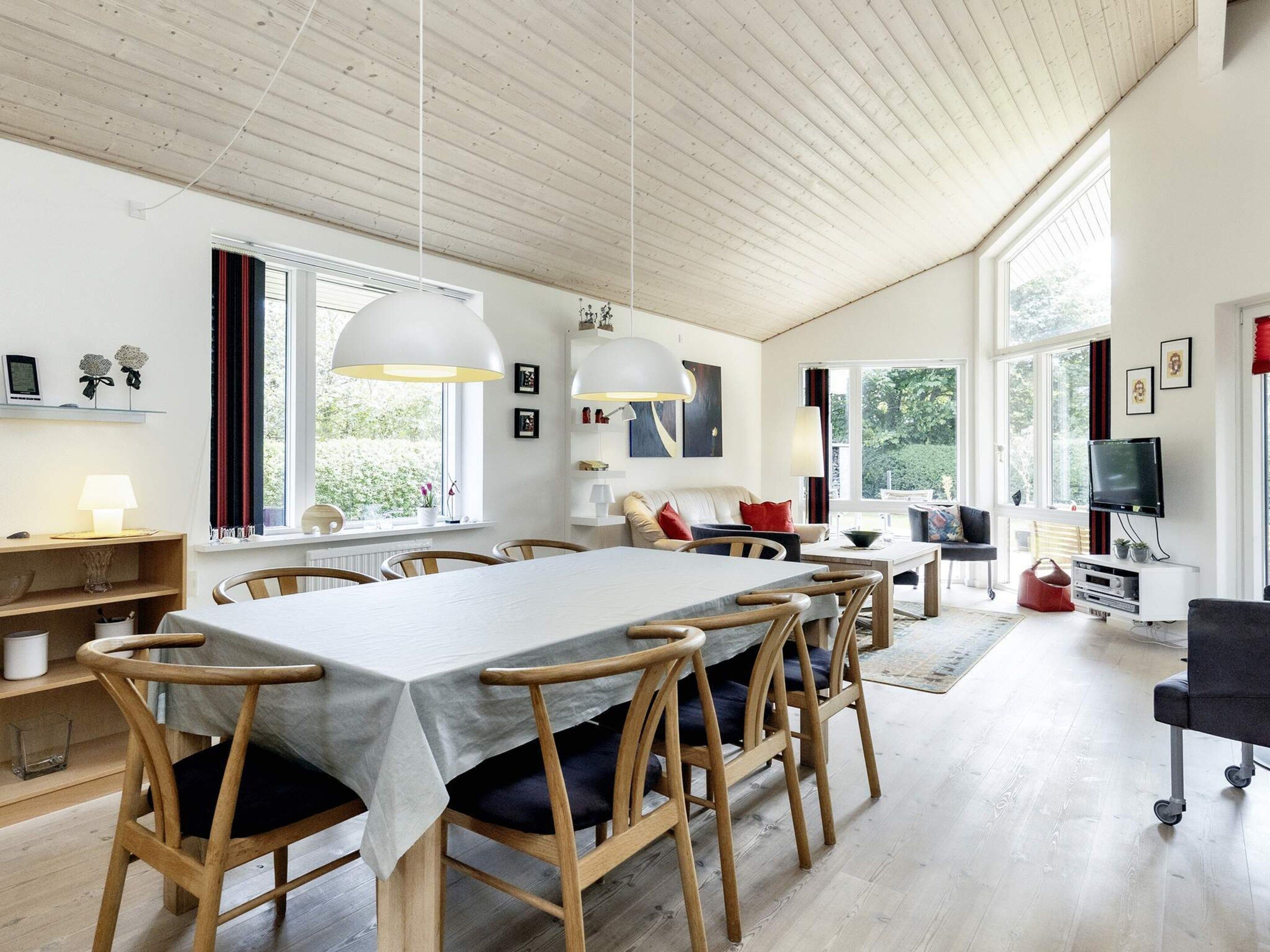 Un grand espace à manger avec une longue table et des chaises en bois, un salon avec un canapé et une télévision, et de grandes fenêtres laissant entrer la lumière naturelle.