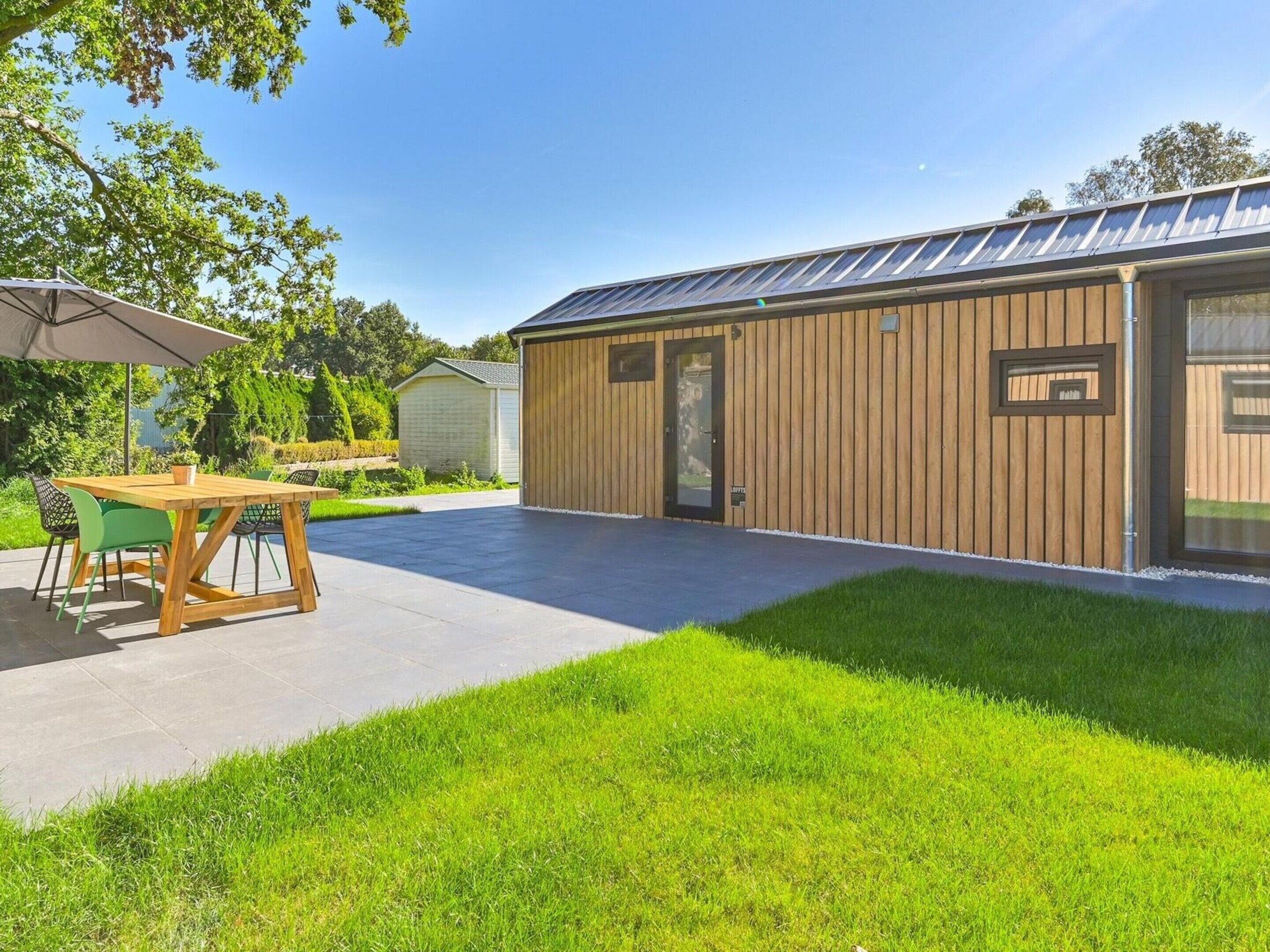 A modern building with a wooden exterior, a covered patio with a table and chairs, and a well-maintained lawn.