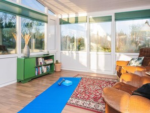 Una sala de estar moderna con ventanas amplias, una estantería verde, un tapete de ejercicios azul, una alfombra estampada y un sofá marrón.