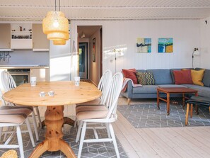 A cozy living area with a round wooden table, white chairs, a grey sofa with colorful cushions, and a patterned rug.