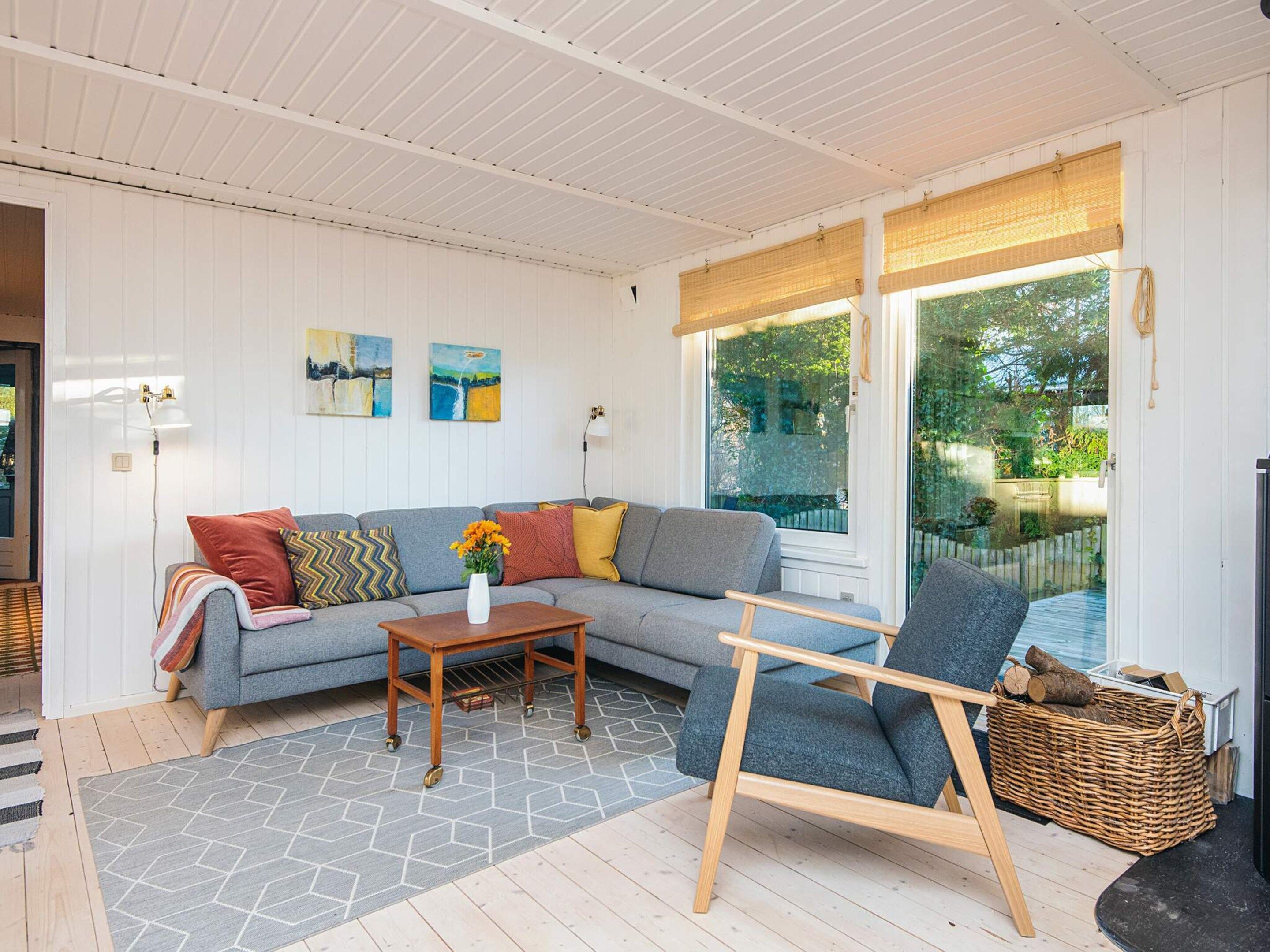 A cozy living room with a grey sofa, a wooden coffee table, and a blue armchair. There are two paintings on the wall, a window with a view of greenery, and a wicker basket with logs.