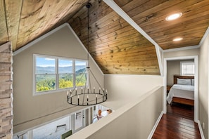 A loft bedroom with a wooden ceiling, a large window with a mountain view, a circular chandelier, and a wooden bed.