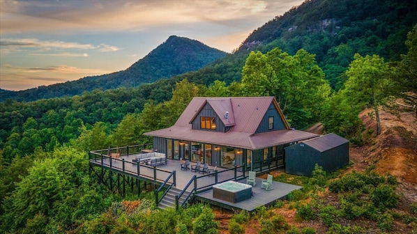 A modern house with a large deck, surrounded by forest and mountains.