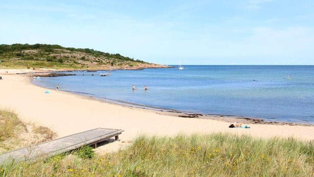 Ein Strand mit einer Holzsteg, ruhigem Wasser und einem Segelboot in der Ferne.