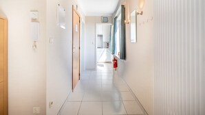 Um corredor limpo e bem iluminado, com chão de azulejos, uma porta de madeira à esquerda e um extintor de incêndio fixado na parede.