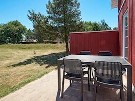 Une terrasse avec une table et des chaises, un bâtiment rouge et une zone herbeuse avec des arbres.