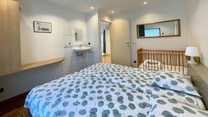 A bedroom with a bed, a wooden crib, a bathroom with a sink and mirror, and a hallway leading to another room. - Villa in Oostduinkerke Near Dunes (Koksijde)