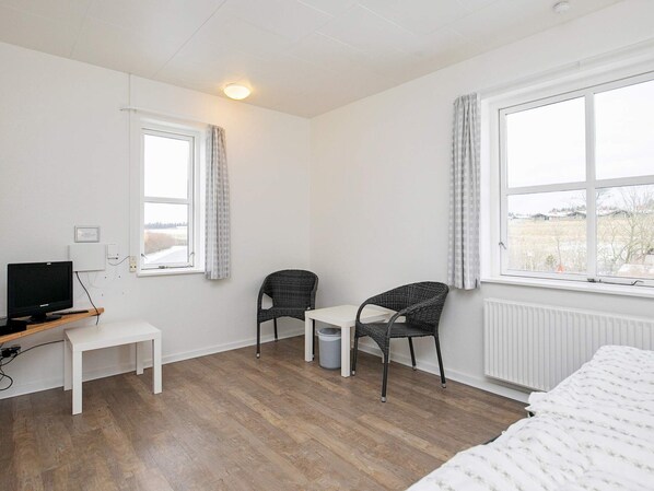 A room with a bed, a desk with a computer, two chairs, a radiator, and large windows with curtains.