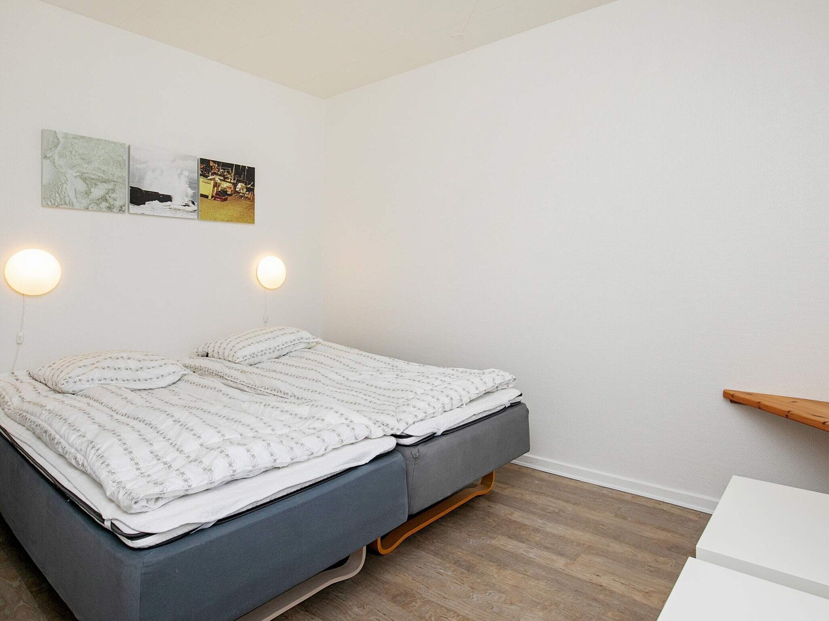 A neatly made bed with a grey mattress and white bedding, two wall-mounted round lights, and a wooden shelf against a white wall.