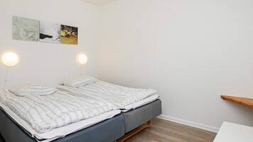 A neatly made bed with a grey mattress and white bedding, two wall-mounted round lights, and a wooden shelf against a white wall.