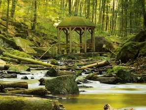A wooden gazebo in a forested area with a stream.