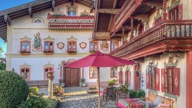Um edifício tradicional com varanda, escadaria e um caminho de pedras que leva a uma área de estar com mesa e cadeiras.