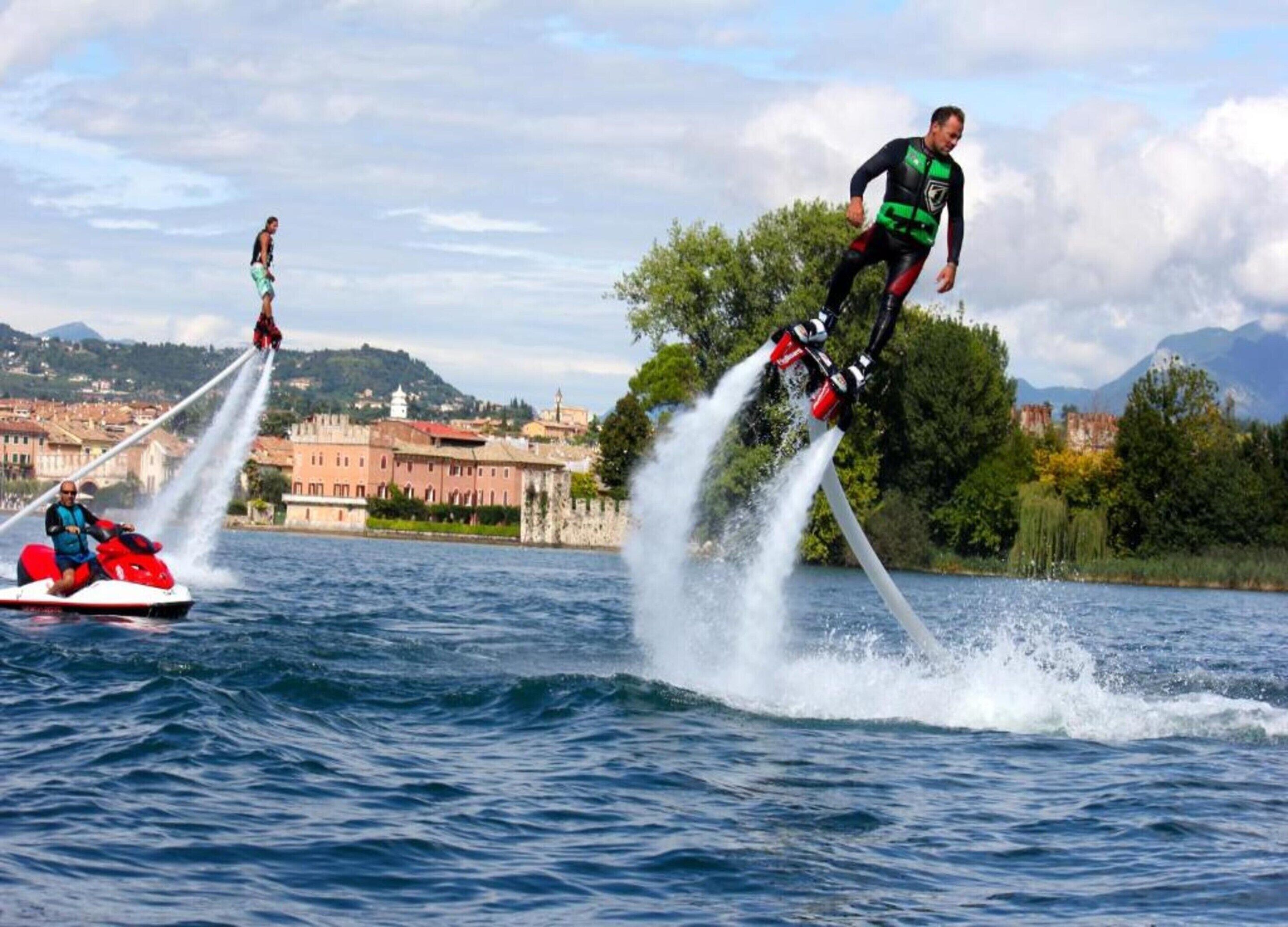 Two individuals jet-skiing on a lake with a cityscape in the background.