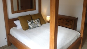 Una cama con dosel de madera, con un cubrecolchón blanco, una mesita de noche con lámpara y un aparador de madera con cajones.