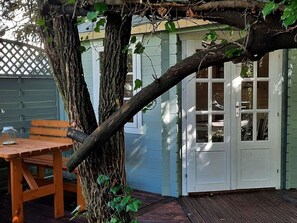 Eine Holzterrasse mit Tisch und Stühlen, ein Baum mit Weinreben und eine weiße Tür mit Fenstern.