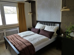 Ein ordentlich bezogenes Bett mit einer braunen Decke, weißer Bettwäsche und zwei Kissen. Zwei Nachttische mit Lampen, ein Fenster mit Vorhängen und eine Heizung sind ebenfalls zu sehen.